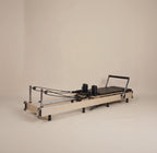 Reformer Pilates con telaio in legno chiaro, carrello imbottito nero e guide metalliche, fotografato su uno sfondo beige.